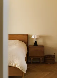 A modern bedroom with a walnut bed frame, white bedding, a bedside table with a unique lamp, and a woven basket.