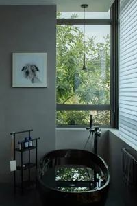 Modern minimalist bathroom featuring a sleek glass tub, grey walls, and a large window showcasing greenery outside.
