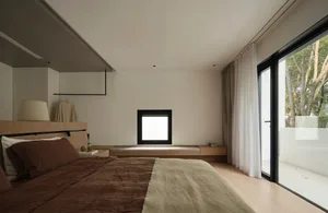 A minimalist bedroom featuring a bed with brown and beige bedding, a wooden headboard, and a window with white curtains.