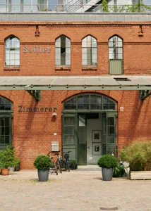 A rustic, industrial building with red brick walls and green window frames, featuring the words 'Sattler Zimmerer' above the 