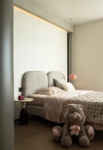 A modern minimalist bedroom featuring a gray upholstered headboard, pink pendant lamp, and a large teddy bear on the floor.