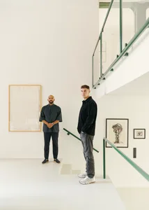Two men standing in a modern art gallery with white walls and minimalist design.
