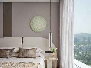 Modern bedroom featuring textured fabric bed, side table with vase and book, and round decorative artwork on the wall.