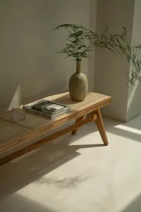 A minimalist living room table featuring natural wood, a woven mat, a beige ceramic vase, and greenery in a corner.
