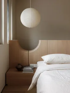 A minimalist bedroom featuring a wooden bed with a pendant light above it, set against a neutral wall and a window with blind