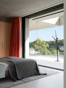 A minimalist bedroom with concrete flooring, a grey blanket, and a large window overlooking green trees.