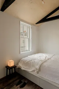 A minimalist bedroom with a white bed, wooden floor, and natural light coming through the window.