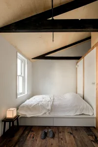 A minimalist bedroom with a white cushioned platform bed placed in a cozy alcove with wooden beams and a small table with a w