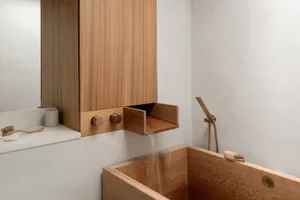 A modern bathroom featuring a wooden bathtub with water flowing out, a wooden cabinet with integrated fixtures, and a minimal