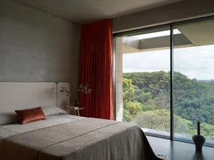 A minimalist bedroom featuring concrete walls and a large window overlooking green foliage.