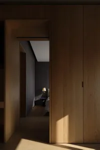A modern, minimalist wooden doorway with a lamp in the adjacent bedroom.