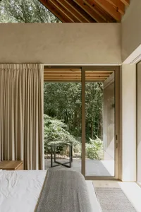 A modern bedroom featuring concrete, glass, and wooden materials, with a view of the forest through large windows.
