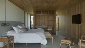 A modern monochromatic bedroom featuring white bedding and a wooden floor with a large bed, wooden shelves, and a wooden chai