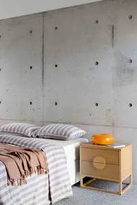 A minimalist bedroom featuring a bed with a concrete headboard and a wooden nightstand, placed against a concrete wall with c