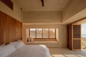 A minimalist bedroom with concrete walls, a bed with white bedding, and cushions on a window seat.