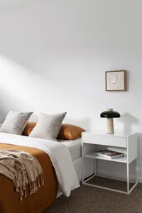 A minimalist bedroom with neutral tones furniture, including a bed with white sheets and orange throw pillows, a white nights