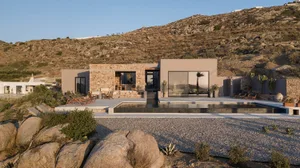 A minimalist modern house with a stucco exterior and stone accents features a sleek, rectangular pool and seating area.