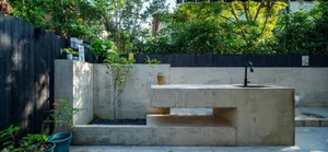 A modern exterior concrete outdoor kitchen with a black faucet and surrounding greenery.