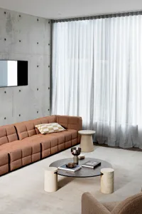 A modern living room with a brown modular sofa, cream-colored marble coffee table, and large windows with sheer curtains.