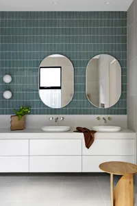 Modern bathroom with green mosaic tile wall, white concrete floating vanity, brass faucets, and a wooden stool.