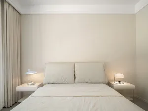 A minimalist bedroom with a bed featuring a simple white frame and soft, neutral-colored linens, illuminated by two bedside l