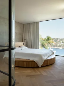 A modern, minimalist bedroom featuring a wooden, round bed with a view of the water.
