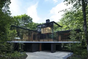 A modern, glass-fronted residential house surrounded by lush green trees and foliage.