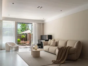A modern living room with a beige sofa, coffee table, and piano, featuring a view of an outdoor space with greenery.