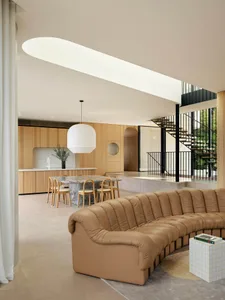 A modern living room features a brown leather sofa, a large white pendant light, and a kitchen area with wooden cabinets and 