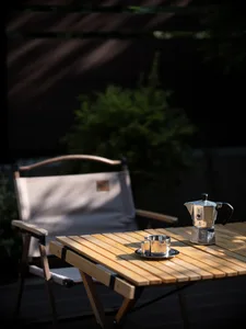 A wooden outdoor coffee table with a coffee pot, mug, and plate, placed outdoors with a folding chair and greenery in the bac