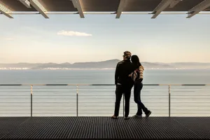 A man and woman standing together on a metallic railing overlooking a coastal landscape.