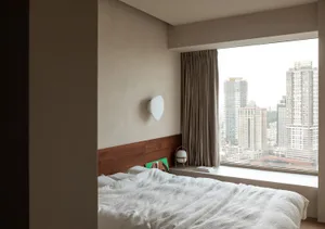 A minimalist bedroom with a large window overlooking the city, featuring a blank white blanket on the bed and a wall sconce.