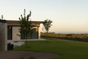 Modern residential exterior with brick walls, glass windows, and a landscaped yard overlooking a forested area during sunset.