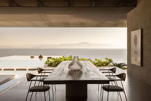 A modern residential interior with a wooden table and concrete chairs overlooking the sea.