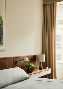 A modern bedroom with a wooden headboard, a bed with a light grey cover, and a nightstand with a white lamp and a green plant