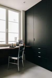 A minimalist office desk setup with a black chair and a window with a white frame, featuring a black cabinet and a wooden sur