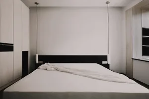 A minimalist bedroom with a white textured bedspread and black headboard against a plain wall with hanging lamps.