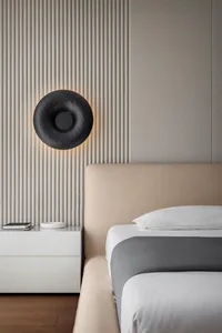 A modern bedroom with a beige upholstered bed, white bedding, and a round black wall sconce on a textured wall.