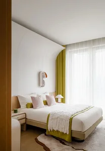 A minimalist bedroom featuring a bed with light-colored bedding and a white, geometric headboard, alongside green curtains an
