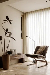 A modern living room with a black leather armchair, white curtains, and a potted plant in a tall vase.