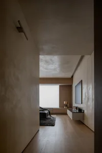 A minimalist bedroom with light wood flooring and furniture, featuring a window with blinds and a carpeted area.