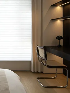 A minimalist workstation featuring a black desk and a woven chair with metal frame, placed by a window with blinds, against a