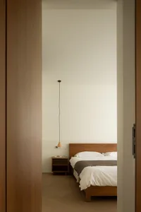 Minimalist bedroom featuring a wooden bed with white bedding and a wall-mounted light fixture against a plain wall.