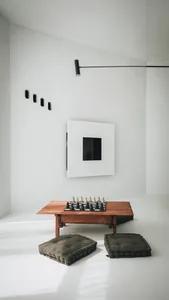 A minimalist living space featuring a wooden table with chess pieces, green cushions, a black wall sconce, and a white wall w