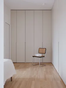 A minimalist bedroom featuring a natural wood floor and a modern chair with a cane seat, against a wall with fitted wardrobes