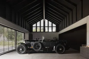 A vintage black car sits in a high-ceilinged, industrial-style garage with concrete walls and floor.