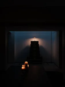 Minimalist interior design featuring stacked dark-colored bags with candles on a table and a lamp above them.