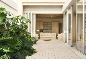 A minimalist living space with wooden furniture and open-plan design, featuring a white brick patio and large green plants.