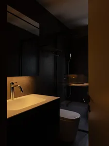 A minimalist black bathroom with a white sink and gold faucet, featuring black tiles on the walls.