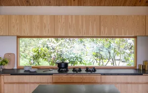A modern, minimalist kitchen with wooden cabinets and a large window overlooking greenery.